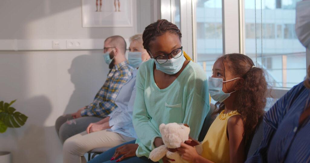 A mother comforts her young daughter as they sit in a medical waiting room. The daughter holds a stuffed bear.