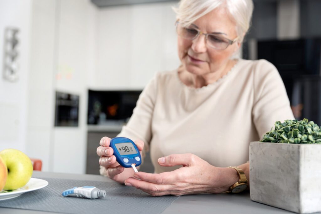 An older woman is sitting at her kitchen table checking her blood sugar with a blood test monitor.